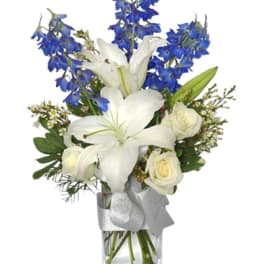 Tall arrangement of white lilies and roses with blue flowers in a clear vase with a silver ribbon.
