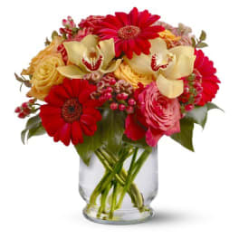 Mixed bouquet of red gerbera daisies, yellow roses, and pale orchids in a clear glass vase