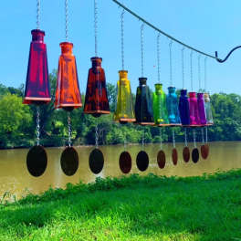 Row of multicolored glass bottle wind chimes hanging from a metal rod outdoors by a river