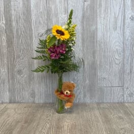 Sunflower and purple floral arrangement in a glass vase with a teddy bear