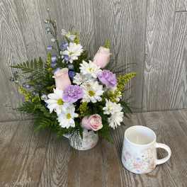 Pink roses and white daisies arranged in a floral mug with a candle beside it.