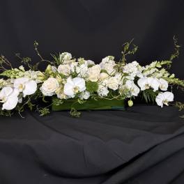 Long white floral arrangement with orchids and roses in a green rectangular container