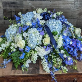 Low blue and white casket spray with hydrangeas, roses, and delphinium accented by navy ribbon.