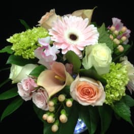 Round arrangement of pale pink gerbera, roses, calla lilies, and green hydrangea in a glass vase