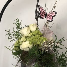 White roses and green blooms in a hanging metal basket with a pink butterfly decoration.