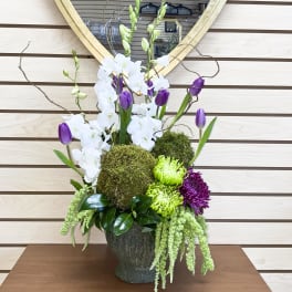 Tall floral arrangement with white orchids, purple tulips, and green moss in a dark vase
