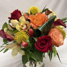 Vibrant arrangement of red and orange roses, yellow pincushion protea, orchids and tulips in a clear glass vase