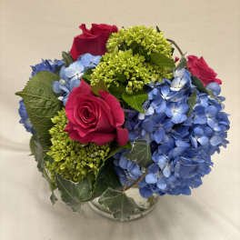 Compact arrangement of hot pink roses, blue hydrangeas, and green blooms in a clear glass vase with ivy.