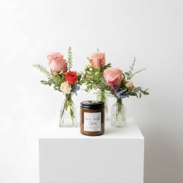 Three small bud vases with pink and red mixed flowers arranged around an amber jar candle on a white pedestal.
