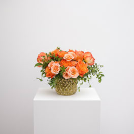 Low arrangement of orange and peach roses in a gold textured glass vase with trailing accents