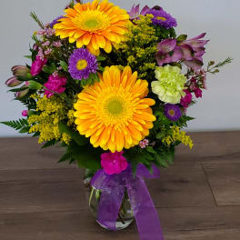 Mixed bouquet of yellow gerbera daisies and purple flowers in a glass vase