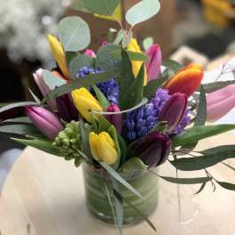 Short glass vase filled with yellow, pink, and purple tulips with purple hyacinths on a light wood table