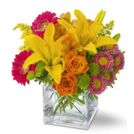 Yellow lilies, orange roses, and pink daisies in a clear square vase