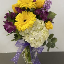 Bouquet of yellow gerbera daisies, roses, purple flowers, and white hydrangea in a glass vase