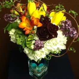 Colorful bouquet with yellow tulips, orange blooms, and dark roses in a glass vase