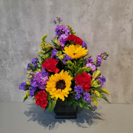Colorful floral arrangement with sunflowers, red carnations, and purple blooms in a black vase