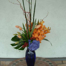 Tall floral arrangement in a blue vase with orange orchids and blue hydrangea