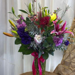 Mixed bouquet with lilies, purple blooms, and a pink ribbon in a glass vase
