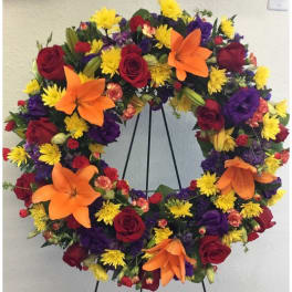 Circular wreath of red roses, orange lilies, yellow daisies, and purple flowers on a stand