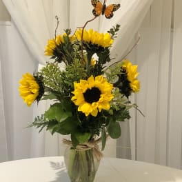 Medium vase of bright yellow sunflowers with accent fillers and a decorative butterfly on curly branches