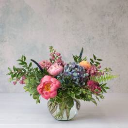 Colorful arrangement of pink peonies, hydrangea, and mixed blooms in a round glass vase