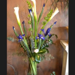 Tall arrangement of white calla lilies, blue irises, and green bells of Ireland in a clear glass vase.