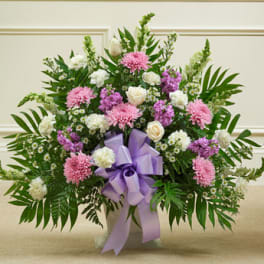 Fan-shaped pink, purple, and white flower arrangement with lavender bow in a white container