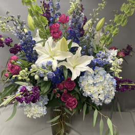 Tall mixed arrangement of white lilies, blue hydrangeas, hot pink roses, and purple blooms in a glass vase