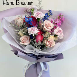 Hand bouquet of pink roses, carnations, and blue flowers wrapped in lavender paper