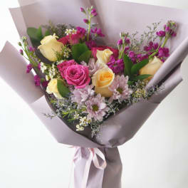 Hand-tied bouquet of yellow and pink roses with purple flowers wrapped in lavender paper.