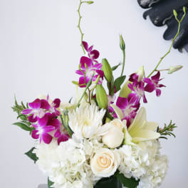 White hydrangeas, roses, lilies, and purple orchids in a green glass cube vase