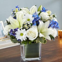 White lilies, roses, and blue flowers in a square glass vase