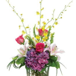 Tall mixed arrangement with pink roses, lilies, purple hydrangea, and yellow orchids in a clear glass vase with stones