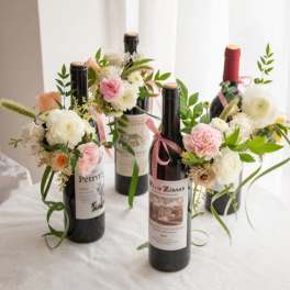 Wine bottles decorated with small pastel pink and white flower clusters and ribbon on a white table.