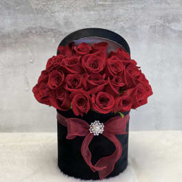 Round black hatbox filled with red roses, tied with a red bow and jeweled brooch.