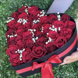 Heart-shaped black box filled with red roses and small white blooms, tied with a red ribbon.