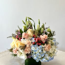 Low mixed arrangement of peach carnations, yellow roses and blue hydrangeas in a dark green glass cube vase