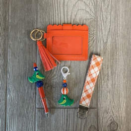 Orange card holder with tassel, pen, keychain, and patterned strap on wood floor