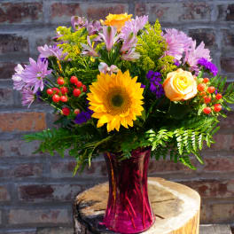 Mixed bouquet with a sunflower, roses, and purple daisies in a pink vase