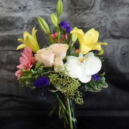 Mixed bouquet with yellow lilies, white orchid, pink blooms, and pale roses in a glass vase