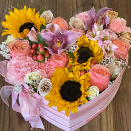 Heart-shaped pink box filled with sunflowers, roses, orchids, and other mixed flowers.