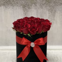 Round black hatbox of red roses wrapped with a red satin bow and jeweled brooch.