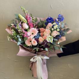 Hand-tied bouquet of pink roses, gerbera daisies, and blue flowers in pink wrap