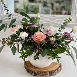 Low pink and white flower arrangement with roses, orchids, and lisianthus in a white bowl on a wood slice base