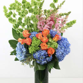 Bouquet of blue hydrangeas, orange roses, and pink snapdragons in a glass vase