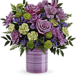 Mixed bouquet of purple roses and blooms with green accents in a lavender striped cylinder vase