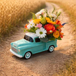 Colorful mixed flowers arranged in the bed of a turquoise ceramic toy truck on a dirt path.