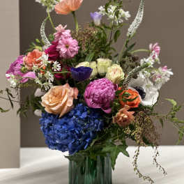 Mixed bouquet in a green glass vase with blue hydrangea and pink, peach, and white blooms