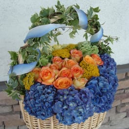 Basket arrangement of orange roses and blue hydrangeas with ivy and ribbon