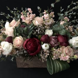 Low wooden box arrangement with blush roses, white ranunculus, and deep red blooms in a lush, elongated design